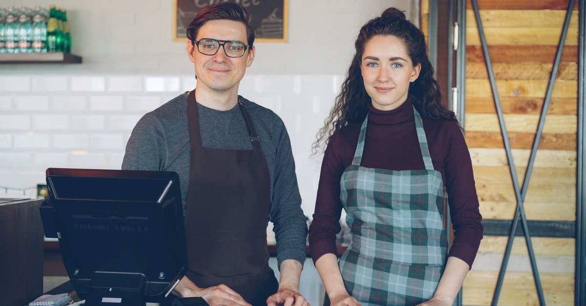 Deux employés de commerce en tablier derrière un comptoir de service, à côté d'un terminal de point de vente
